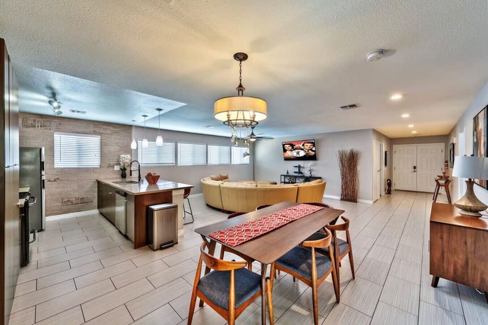 The open-plan living area features a dining space with a wooden table, six blue-cushioned chairs, and a chandelier. A curved beige sectional faces a wall-mounted 65` Roku TV. The kitchen with an island and pendant lights is also in view.