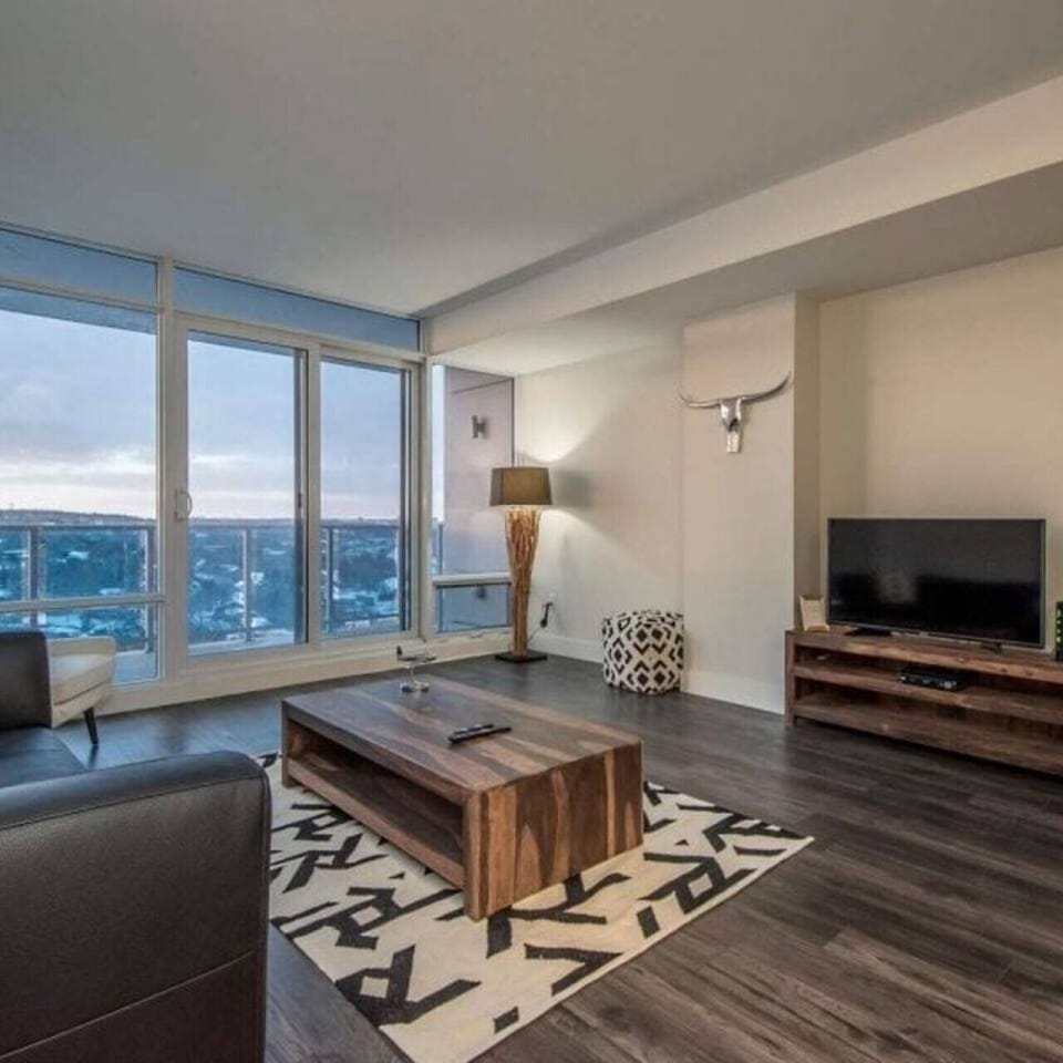 Relax in this spacious living room featuring floor-to-ceiling windows with expansive city views, rich dark wood furnishings, a flat-screen TV, and modern Southwestern-inspired decor.