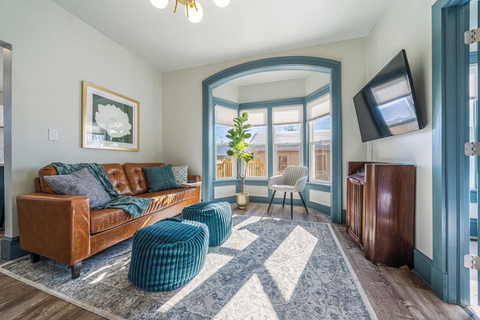 - Bright living room with bay windows filling the space with natural light   - Modern leather sofa and plush ottomans for relaxing evenings   - Smart TV setup perfect for streaming and cozy gatherings