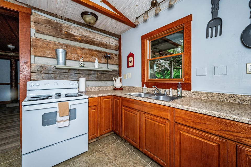 Kitchen features natural wood cabinets, granite-patterned countertops, and a white electric stove beneath a rustic log accent wall.