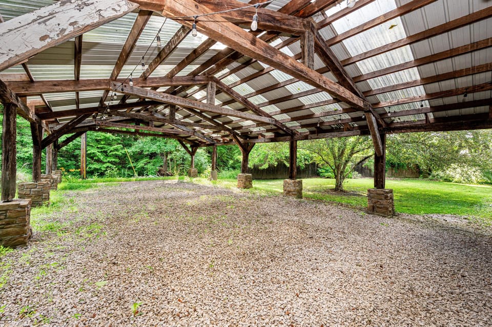 Open-air wooden pavilion with stone pillars, gravel floor, and translucent corrugated roof panels overlooking a green yard.