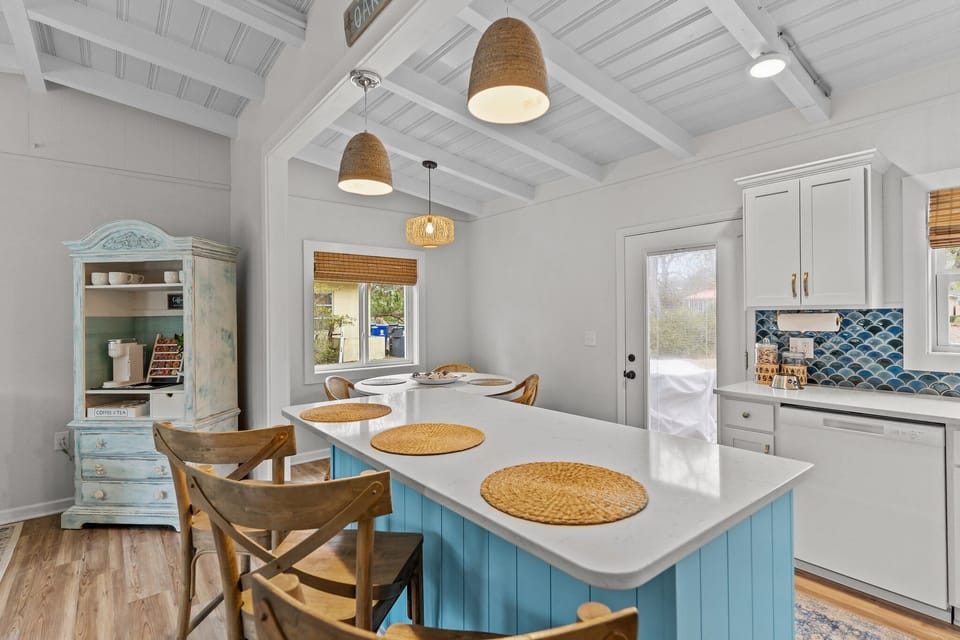 Bright open kitchen and dining area in Oak Island NC featuring a large blue island with seating, coastal lighting, round dining table with wicker chairs, and views into the comfortable living room.