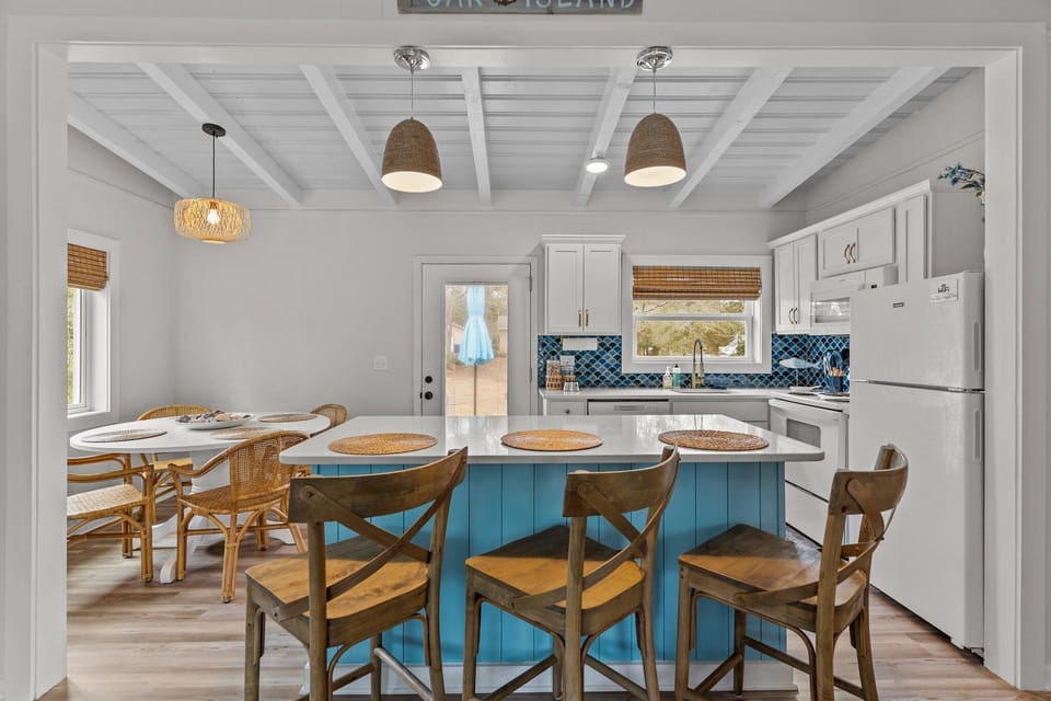 Bright open kitchen and dining area in Oak Island NC featuring a large blue island with seating, coastal lighting, round dining table with wicker chairs, and views into the comfortable living room.