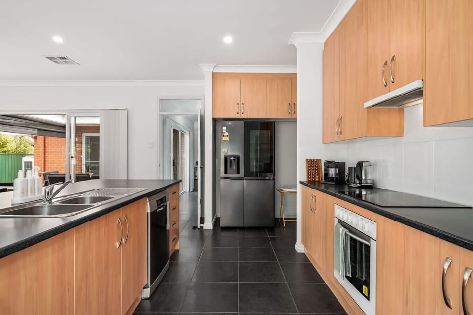 A modern kitchen with light wood cabinetry and black countertops. It features a stainless steel refrigerator, a sink, a dishwasher, and an oven. The kitchen has a sleek design with dark tiled flooring and is well-lit with recessed ceiling lights.