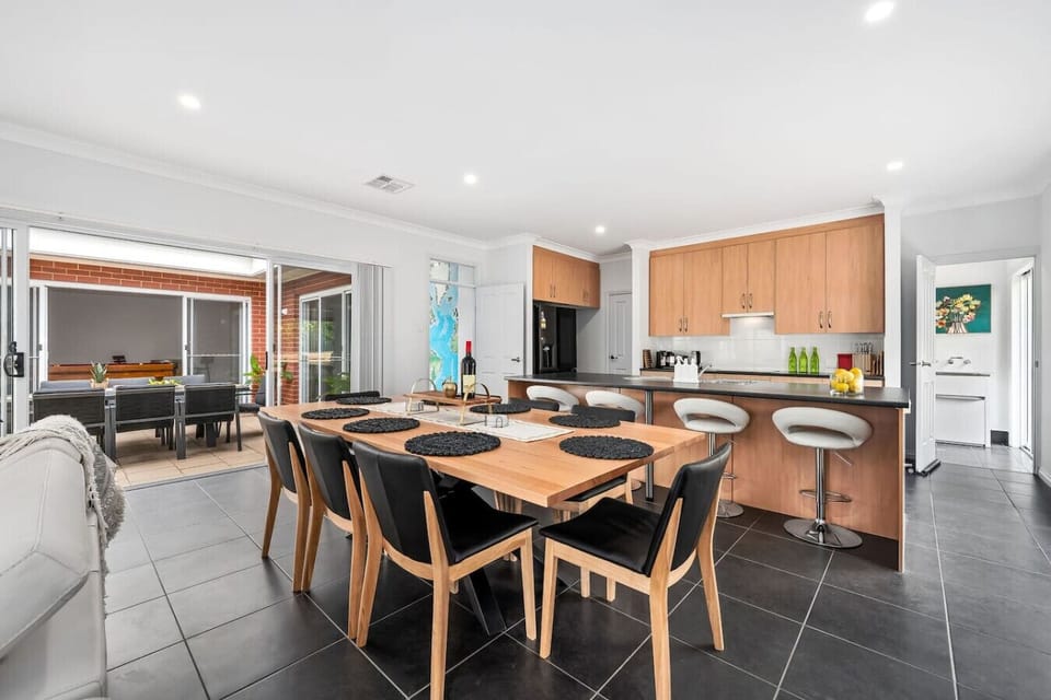 A modern, open-plan kitchen and dining area with a sleek design. It features a wooden dining table surrounded by black and wooden chairs, a kitchen with light wooden cabinets, and a view of an outdoor patio area through large sliding doors. The space is w