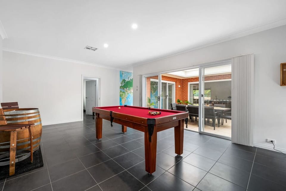 A spacious room with a dark tiled floor, featuring a pool table with a red top as the central element. There are also wooden barrel stools on the left side, and large sliding doors in the background that open to an outdoor area with a dining set, allowing