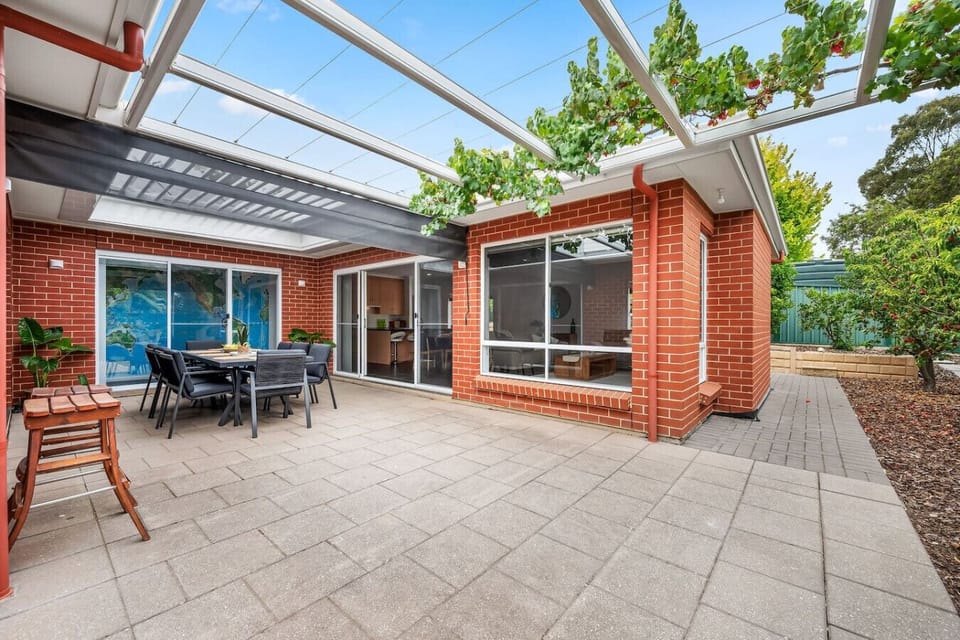 A charming outdoor patio area adjacent to a brick house with large glass doors. The patio features a set of outdoor furniture, including a table and chairs, and is covered by a modern pergola with glass panels and some climbing plants adding a touch of gr