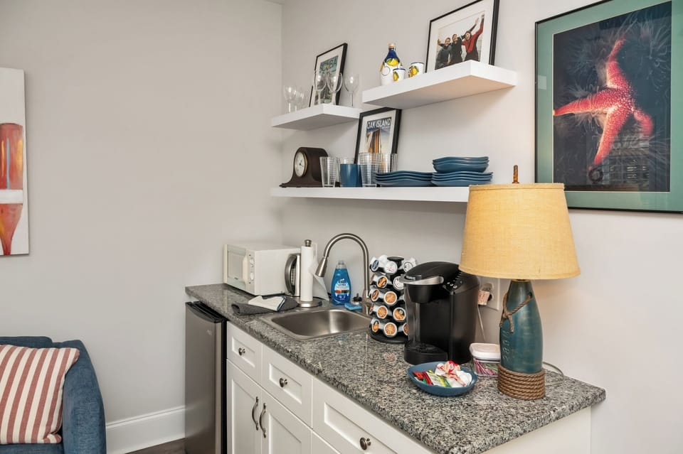 Kitchenette with refrigerator, microwave, sink, kettle, glassware and utensils. 