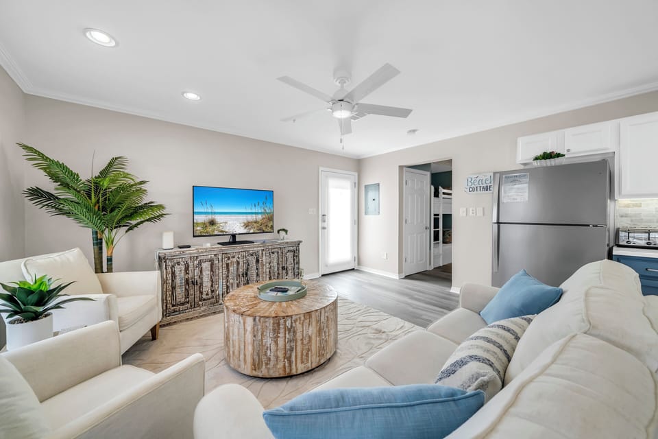 Unwind in your bright, coastal-inspired living room with plush seating, a large flat-screen TV, and breezy ceiling fan. Natural textures and fresh greenery create a relaxing beach getaway atmosphere.