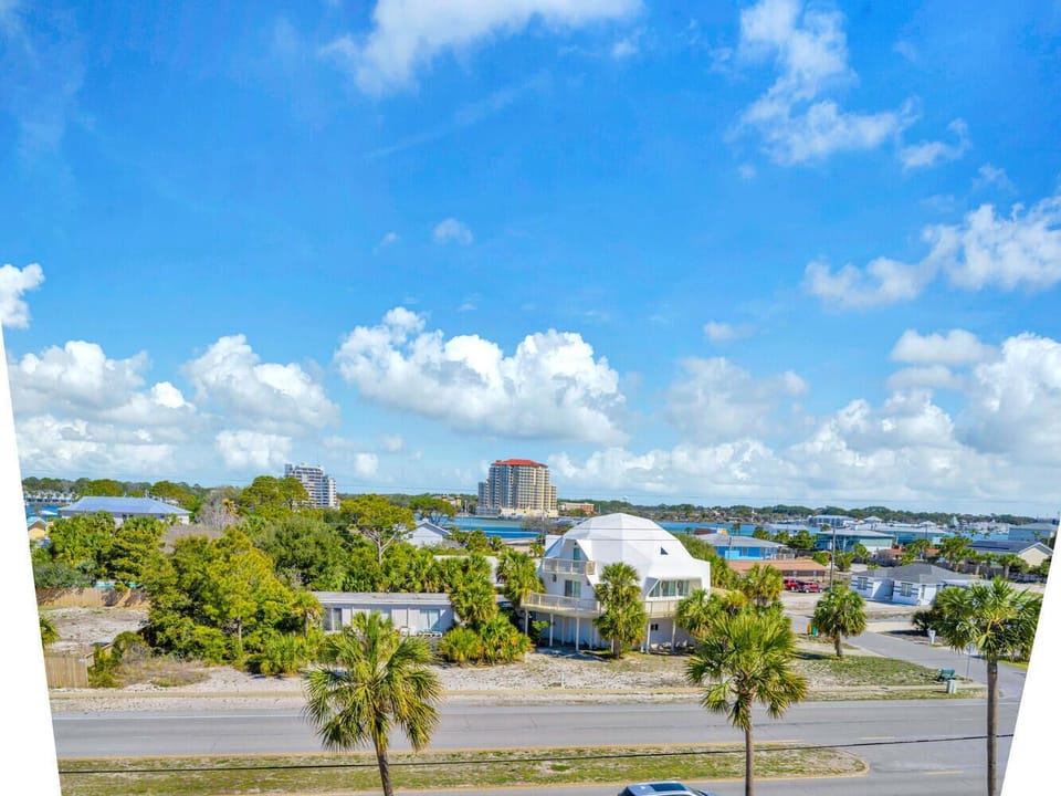 Aerial view of coastal neighborhood with palm trees, residential buildings, and nearby high-rise developments under bright blue skies.
