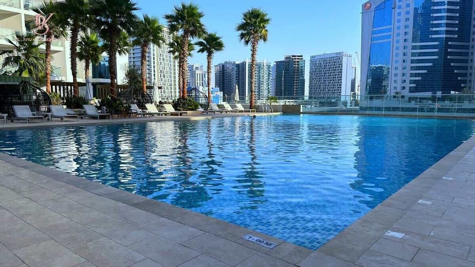 A sparkling swimming pool glistens under sunlight, its clear blue water reflecting the sky. Smooth tiles surround the pool, with a few loungers and palm plants nearby, creating a serene, inviting, and resort-like atmosphere.