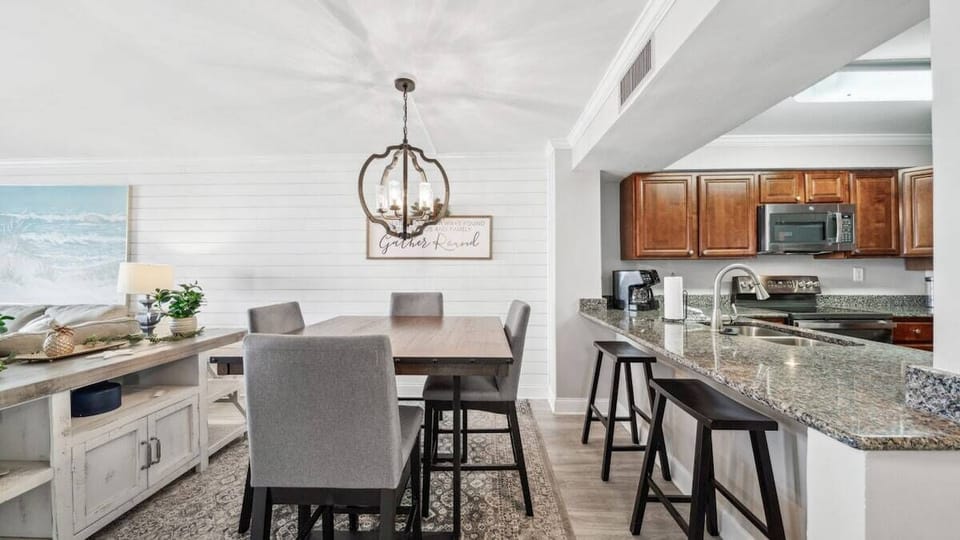The dining area offers a chic wooden table with upholstered seats, a statement chandelier, and nearby bar seating connecting to the kitchen.
