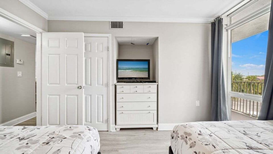 This cozy guest bedroom has two comfortable beds, a wall-mounted TV above a white dresser, and natural light from a balcony-facing window.