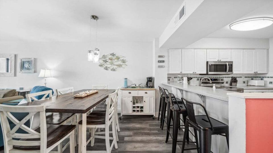 The open dining area features a rustic farmhouse table and breakfast bar seating, perfect for family meals or casual gatherings. Coastal decor and bright finishes create a welcoming space for everyone to enjoy.