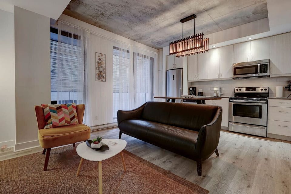 A bright and welcoming space with an industrial-chic touch, combining a modern kitchen, cozy seating, and natural light streaming through large windows.