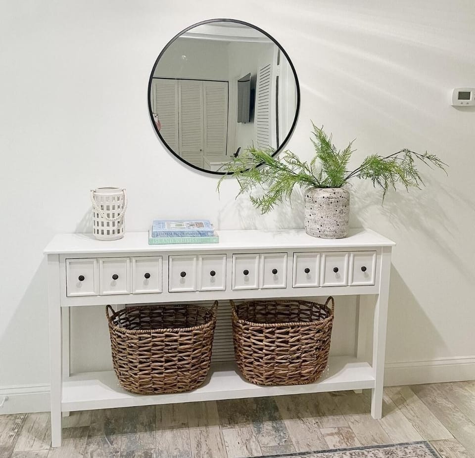 Entryway console with nine small drawers, two woven baskets on the lower shelf, round black-framed mirror, and decorative vase.