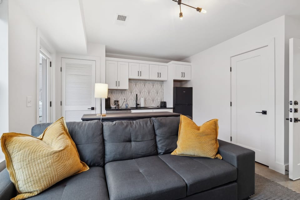Living Room: Seamless blend of living and kitchen spaces with a cozy sectional, mustard accents, and a fully equipped kitchen.