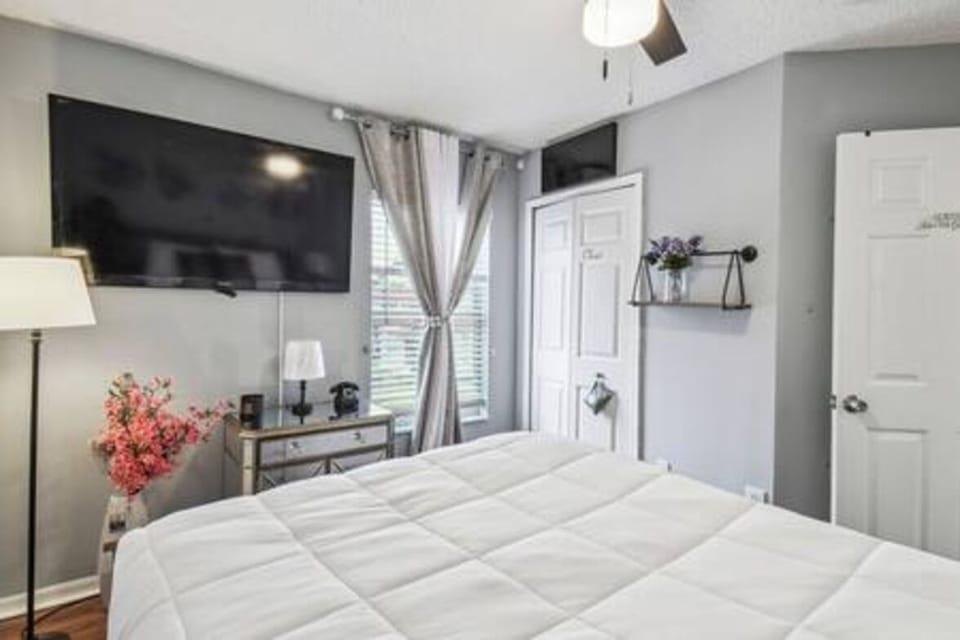 Enjoy comfort in this inviting bedroom with a plush white bedspread and a wall-mounted TV for entertainment. Natural light pours through chic curtains, highlighting the soothing gray tones and cozy decor for a relaxing atmosphere.