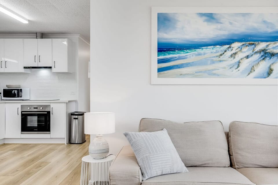 A modern, minimalist living space with a light color scheme. It features a beige sofa with a light-colored pillow, a small side table with a decorative lamp, and a white kitchen area with cabinets, an oven, and a microwave. A large, striking painting of a