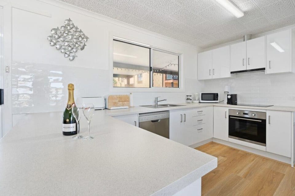 A modern kitchen with white cabinets, a light-colored countertop, and wooden flooring. There is a bottle of champagne and two glasses on the countertop, suggesting a celebratory or special occasion. The kitchen also features a window, a microwave, a stove