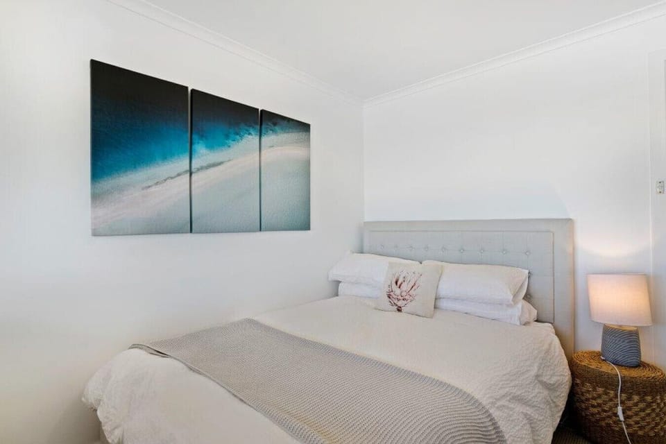 A neatly arranged bedroom with a light-colored bed adorned with white pillows and a decorative cushion. A modern three-panel artwork with a beach scene hangs on the wall above the bed. Next to the bed, there is a woven bedside table with a table lamp, cre
