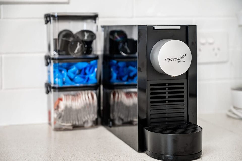 A black espresso machine labeled `espresso system` placed on a kitchen countertop. Behind it, there are clear storage containers holding coffee pods, some of which are visible through the transparent sides of the containers. The setting appears to be a cl