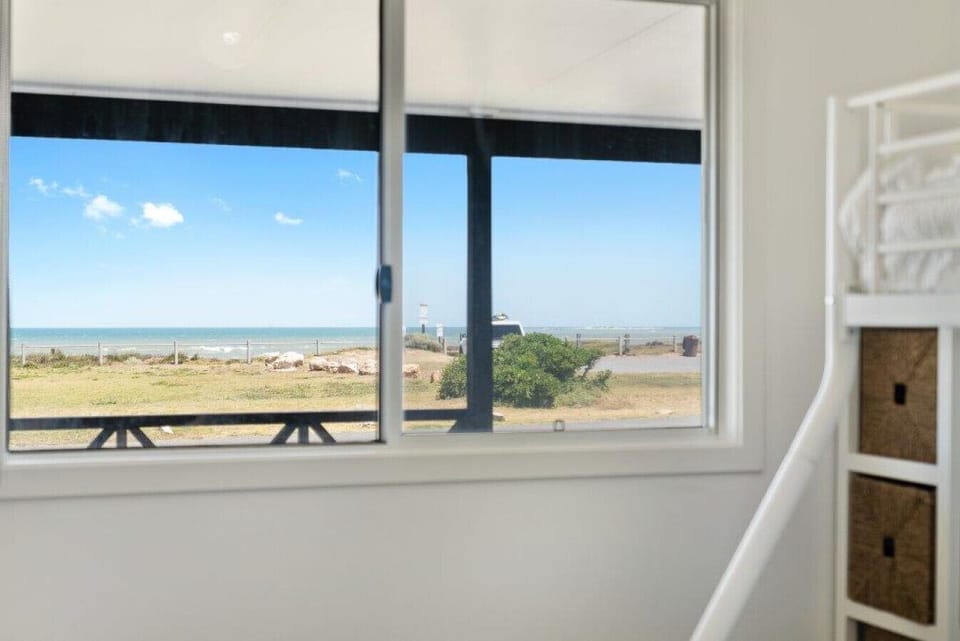 A view from inside a room through a window, where the outside scene includes a grassy area, some bushes, and the ocean in the distance under a clear blue sky with a few clouds. Inside the room, there is a white bunk bed with storage drawers underneath.