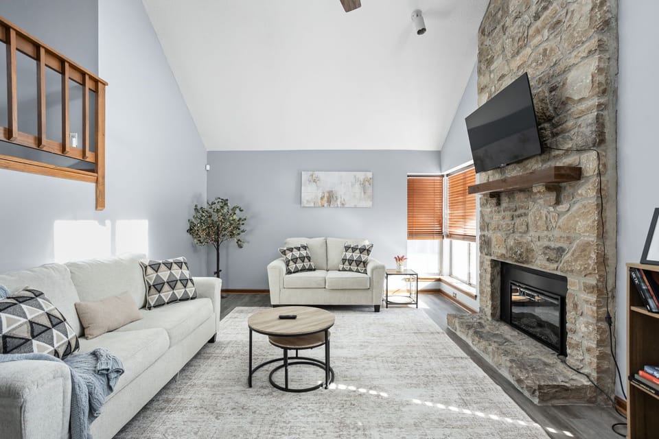 The main living area features two couches, a flat screen TV, electric fireplace, a bookshelf with books, and plenty of light!
