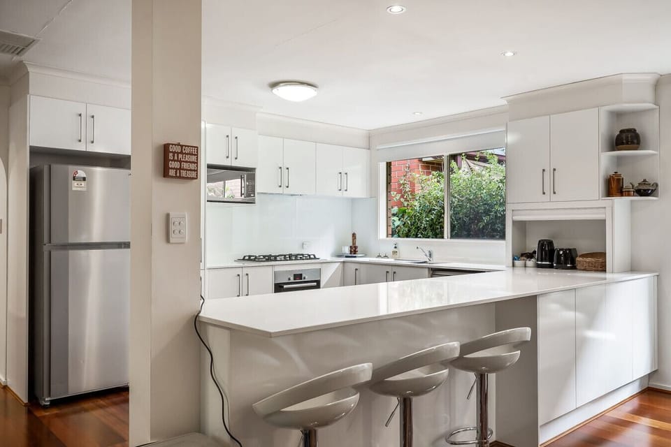A modern kitchen with white cabinetry and a large white countertop. There are several stools in front of the counter, a stainless steel refrigerator, and a view of greenery outside the window. The kitchen has a clean and bright appearance with wooden floo