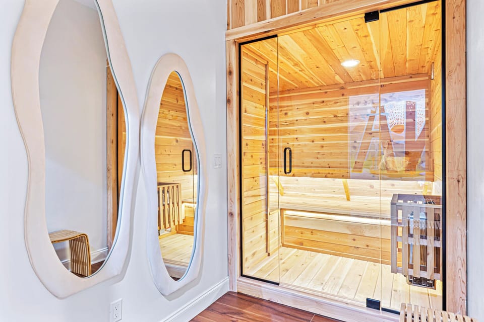 A beautifully designed home sauna with warm wooden interiors and a sleek glass door offers a relaxing retreat. Outside the sauna, two artistic wavy mirrors add a stylish touch to the modern, serene space.