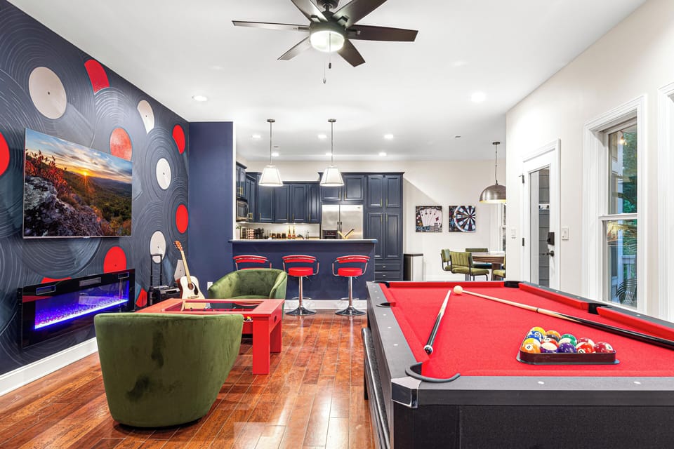 A vibrant entertainment space featuring a red-felt pool table, a cozy seating area with green velvet chairs, and a modern electric fireplace beneath a mounted TV. The stylish bar area, adorned with navy blue cabinetry and red barstools, seamlessly connects to a game corner with a dartboard and poker artwork, creating the ultimate game room experience.