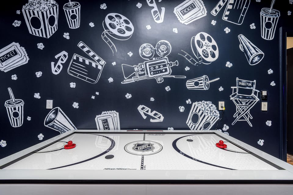 A sleek NHL-branded air hockey table sits in front of a striking black-and-white cinematic mural, featuring popcorn, film reels, director’s chairs, and other movie-themed elements. This space combines arcade fun with a stylish theater ambiance, perfect for entertainment lovers.