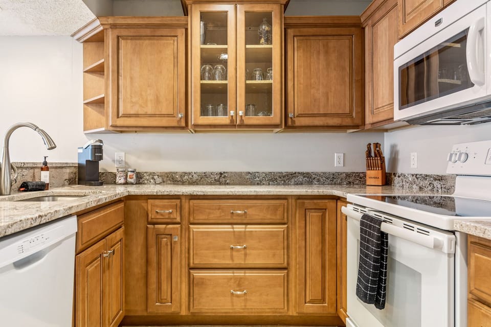 Kitchen angle showing wood cabinetry, counters, sink, dishwasher, range, microwave, coffee maker, and glassware storage.