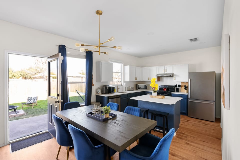 Modern dining area with a stylish wood table, plush blue chairs, and a chic open kitchen. Perfect for family meals after a day at Dell Diamond or Kalahari! The open layout leads to a private backyard, great for relaxing or enjoying the Texas sunshine