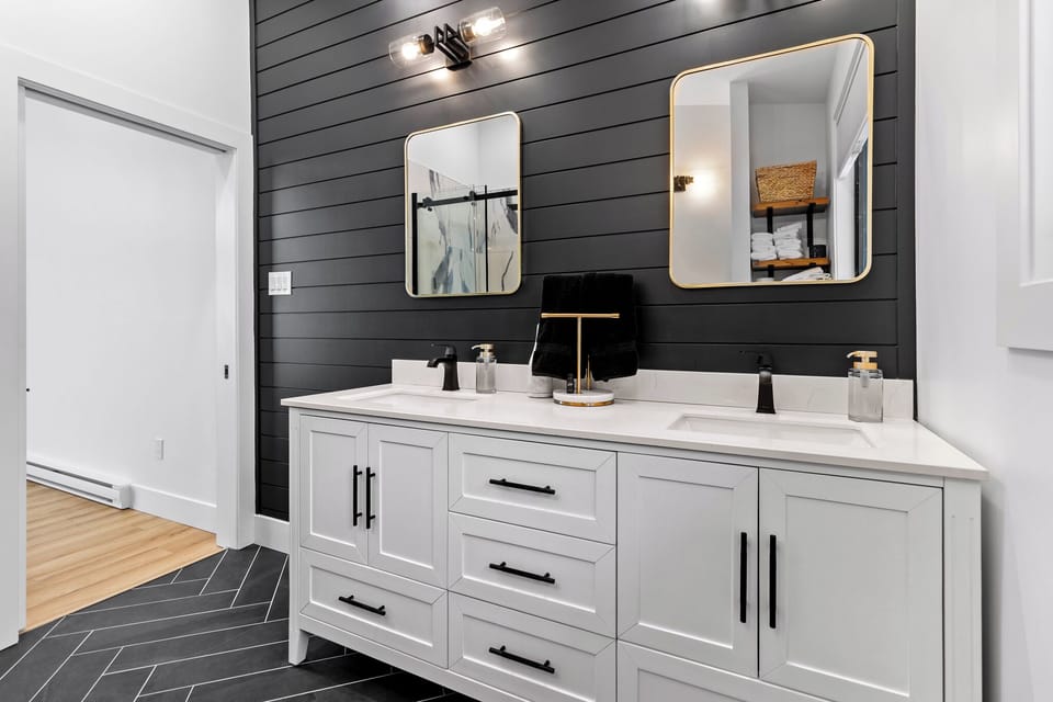 Modern bathroom with double sinks, black fixtures, white cabinetry, black handles, gold-framed mirrors, and a black shiplap accent wall. Contemporary and sleek design.