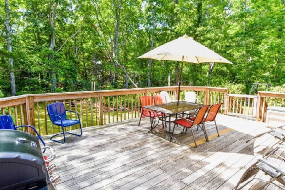 Spacious back deck with a grill, dining table, and umbrella—perfect for enjoying a meal after a relaxing day at the beach, surrounded by lush greenery.
