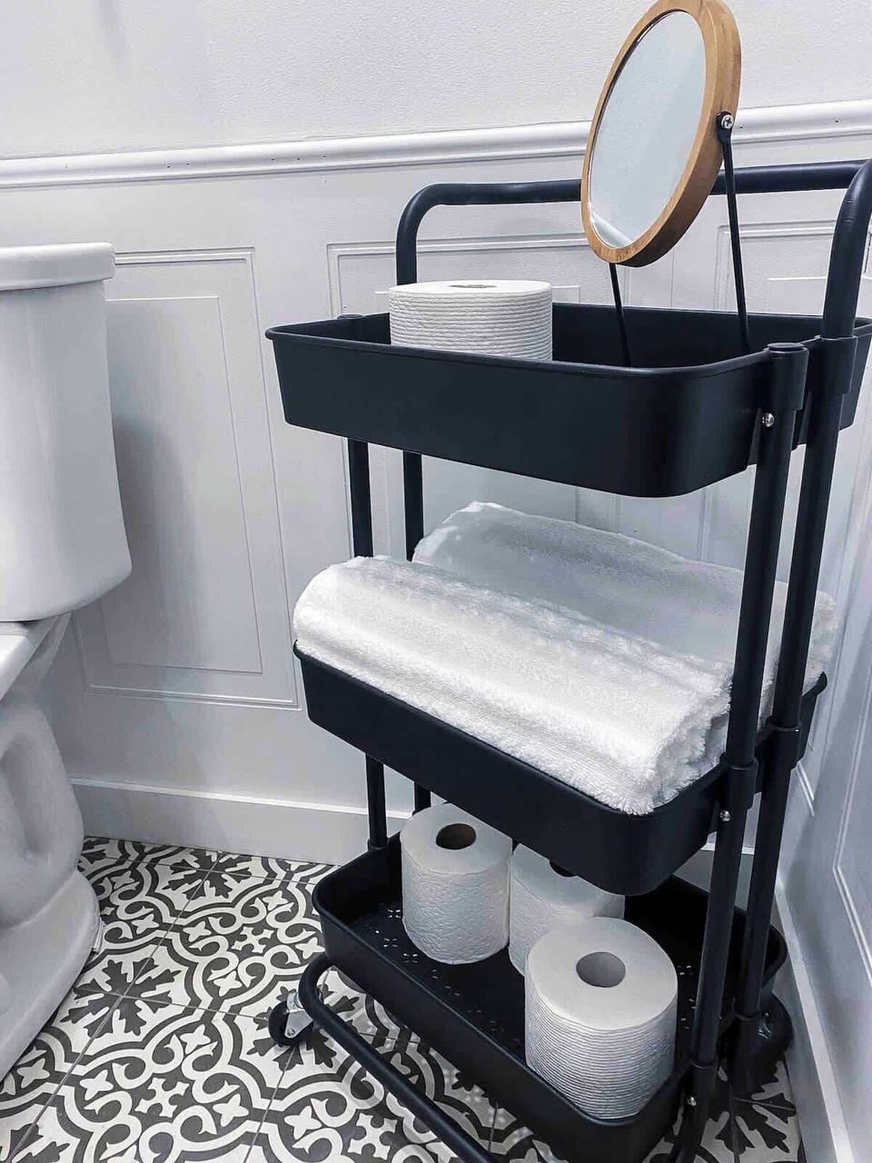 Refresh and prepare with ease using this black three-tiered storage cart holding bath essentials, extra toilet paper, rolled towels, and a small round mirror, set over decorative black-and-white floor tile.