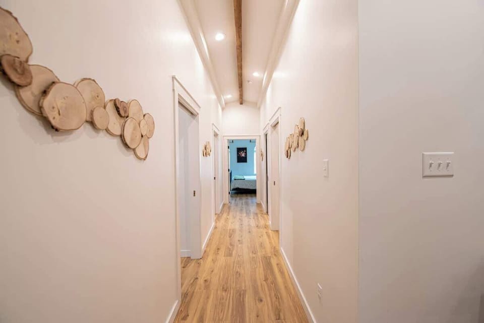 Walk down this bright, airy hallway featuring light wood-look flooring, exposed ceiling beams, recessed lighting, and charming natural wood slice wall art accents guiding the way to adjacent rooms.