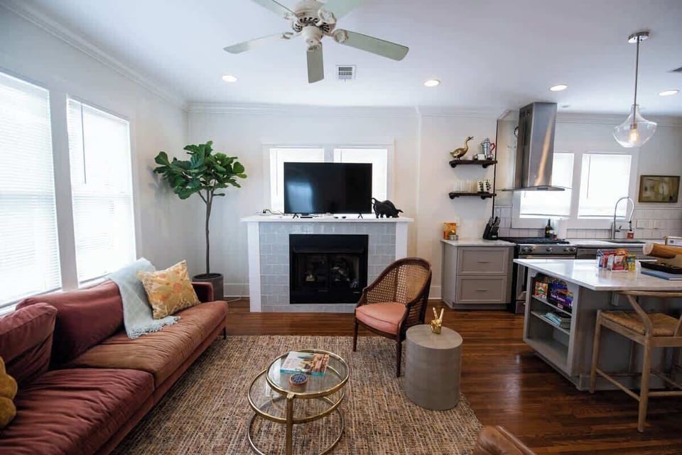Relax in this airy, open-concept space featuring a cozy living room with a rust-orange sofa, woven accent chair, glass coffee table, and a fireplace topped with a television, adjacent to a modern kitchen with a large island setup.