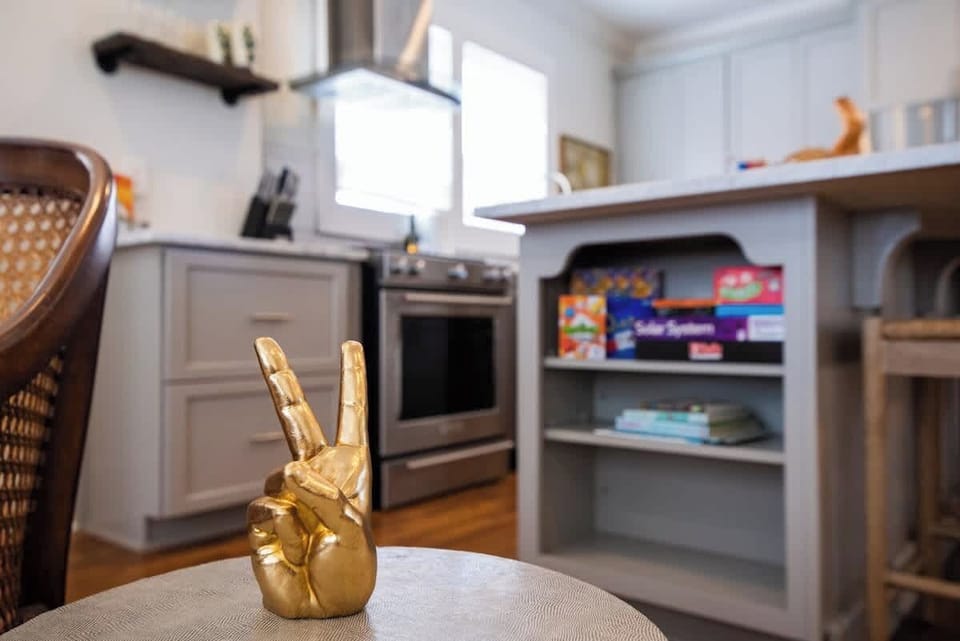 Accent your space with this stylish kitchen corner featuring a gold peace sign sculpture on a small side table, positioned in front of a grey kitchen island with open storage shelves for games and books, along with stainless steel appliances.