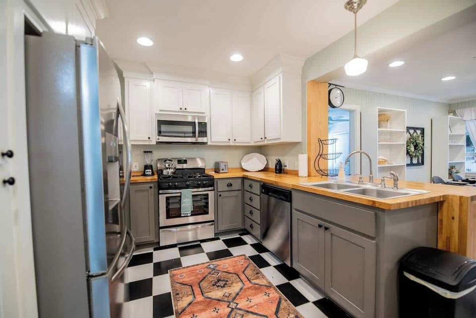 Cook with ease in this charming kitchen, featuring grey lower cabinetry paired with white upper cabinets, butcher block countertops, stainless steel appliances, classic black-and-white checkered floor tile, and a patterned runner rug.