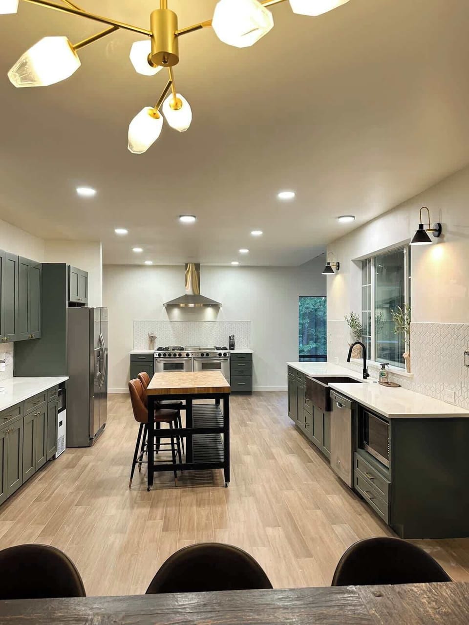 Gather and cook in this modern, expansive kitchen featuring olive green cabinetry, light wood-look flooring, a butcher block island with seating, stainless steel appliances, and a stylish brass chandelier overhead.