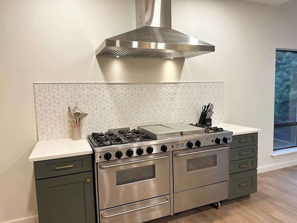 Cook with ease in this sleek kitchen set-up featuring a large stainless steel double-oven range, a matching vent hood, white geometric geometric tile backsplash, and dark olive green cabinetry.
