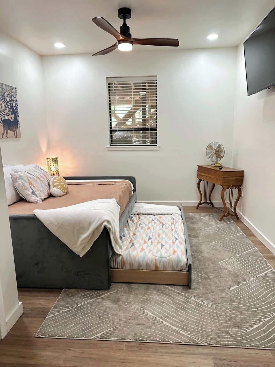 Relax in this versatile bedroom featuring a dark grey daybed with a pulled-out trundle bed, a wall-mounted TV, a small wooden console table, a ceiling fan, and a neutral geometric rug.