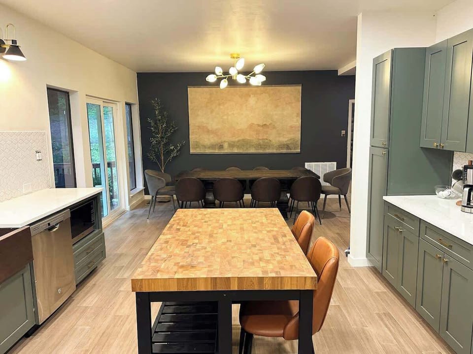 Prepare meals in this spacious, open-concept kitchen and dining area, featuring sage green cabinetry, a butcher block island with leather barstools, and a dining table set against a dark accent wall with map art.