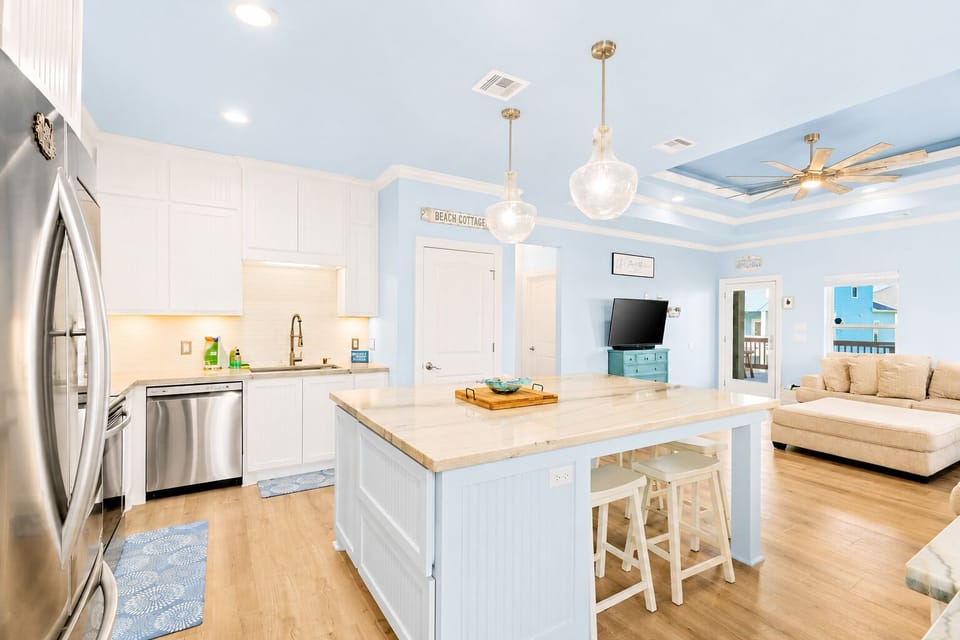 Coastal kitchen with a large island, white cabinetry, stainless steel dishwasher and refrigerator, pendant lighting and an open view into the living room.