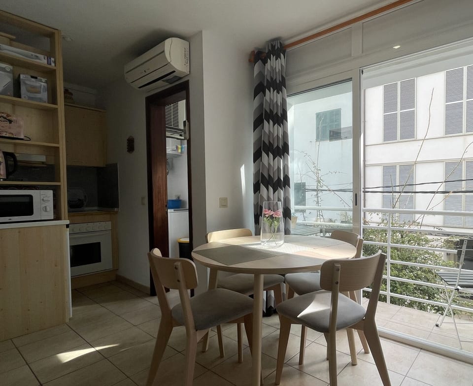 Bright dining area with balcony access, wooden table with chairs, and an integrated kitchen. Cozy space with air conditioning and large windows."