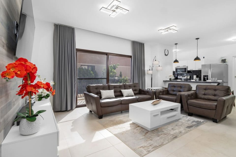 Unwind in this stylish living room, featuring comfy leather seating, a modern coffee table, and a vibrant floral accent. Large windows bring in natural light, connecting the cozy space to the kitchen area