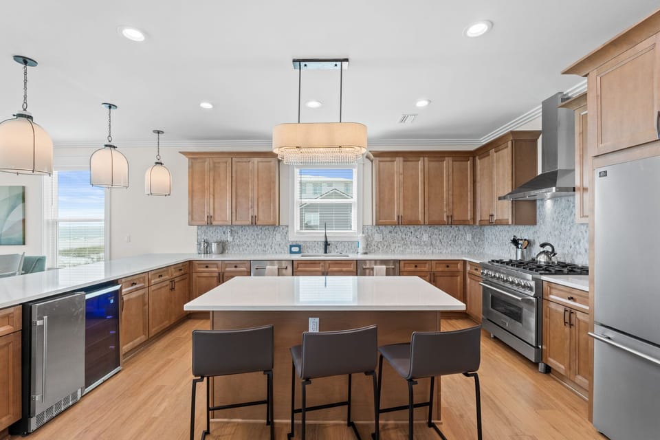 Your vacation kitchen features a spacious island for gathering, modern appliances, warm wood cabinetry, and natural light streaming through windows with ocean views.
