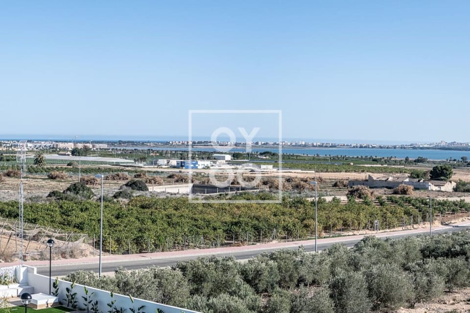 Enjoy stunning panoramic views from this balcony: lush orchards and olive groves stretch below, leading to tranquil lagoon waters and distant city skyline, all under a clear blue sky. 🌳🌊🏙️☀️